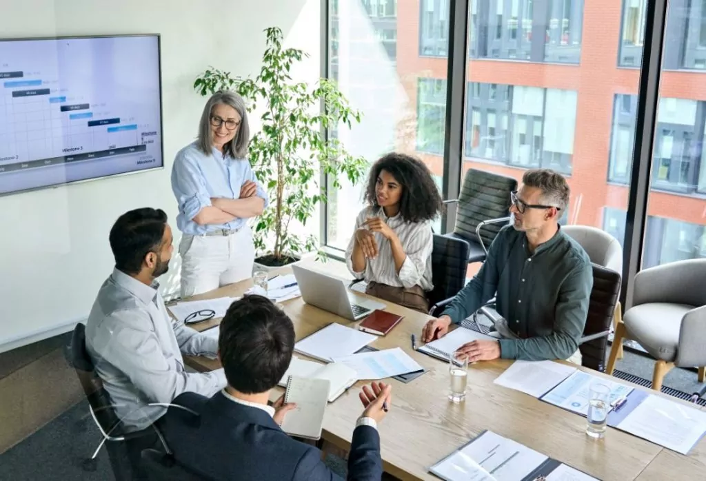 Four senior executives attending leadership training in a board room with the trainer standing at the head of the table where the others are sitting explaining what makes a good leader. 