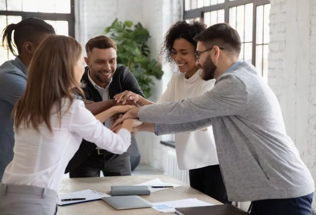 Five employees huddled together with their hands over one another's as their workplace experiences improved team engagement and performance. 