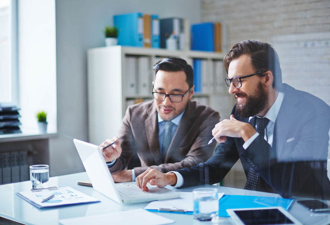 Two business professionals in suits collaborating at a laptop during a leadership training session, discussing strategies and problem-solving in a modern office setting.